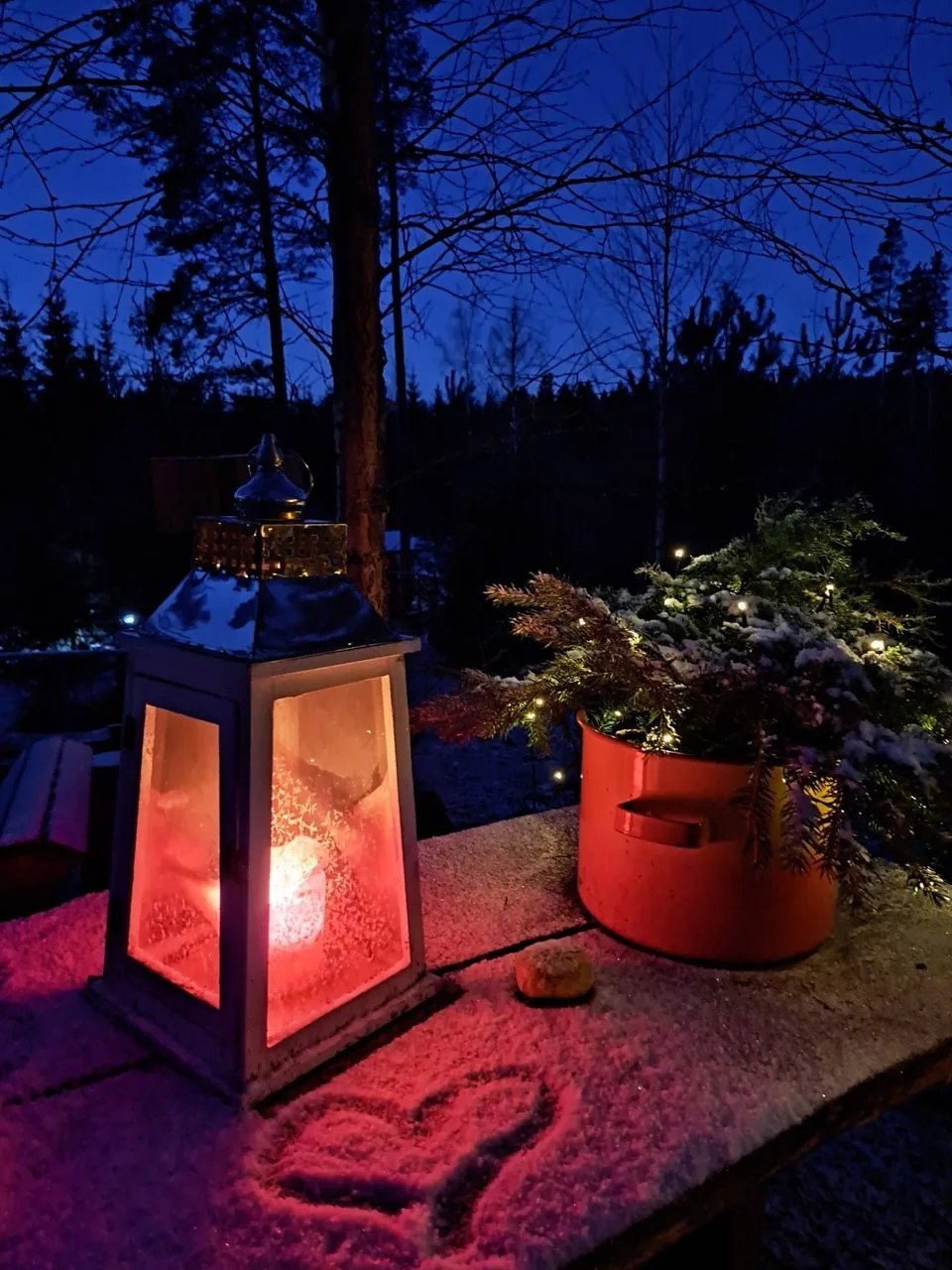 Lyhty palaa lumisella pöydällä illan sinisessä hämärässä metsän laidalla